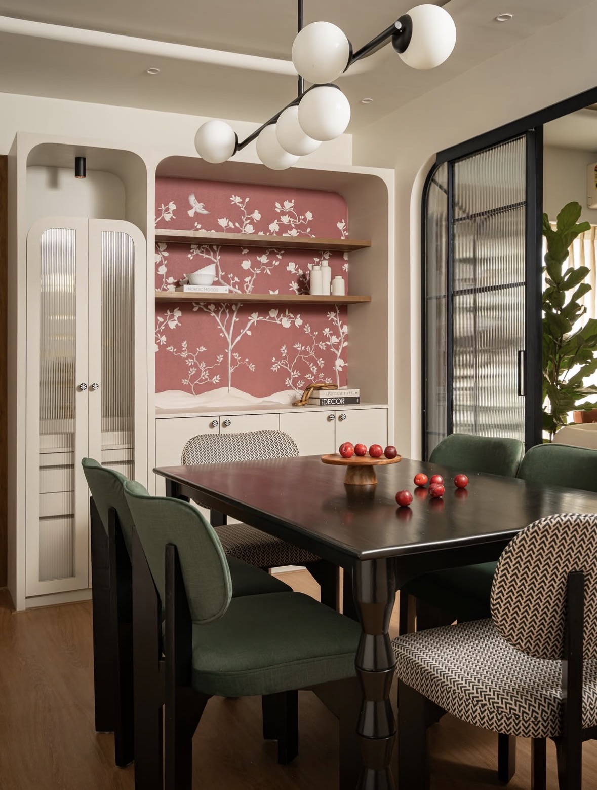 Dining area with a black table, green and patterned chairs, a globe pendant and a rounded niche with pink floral wallpaper