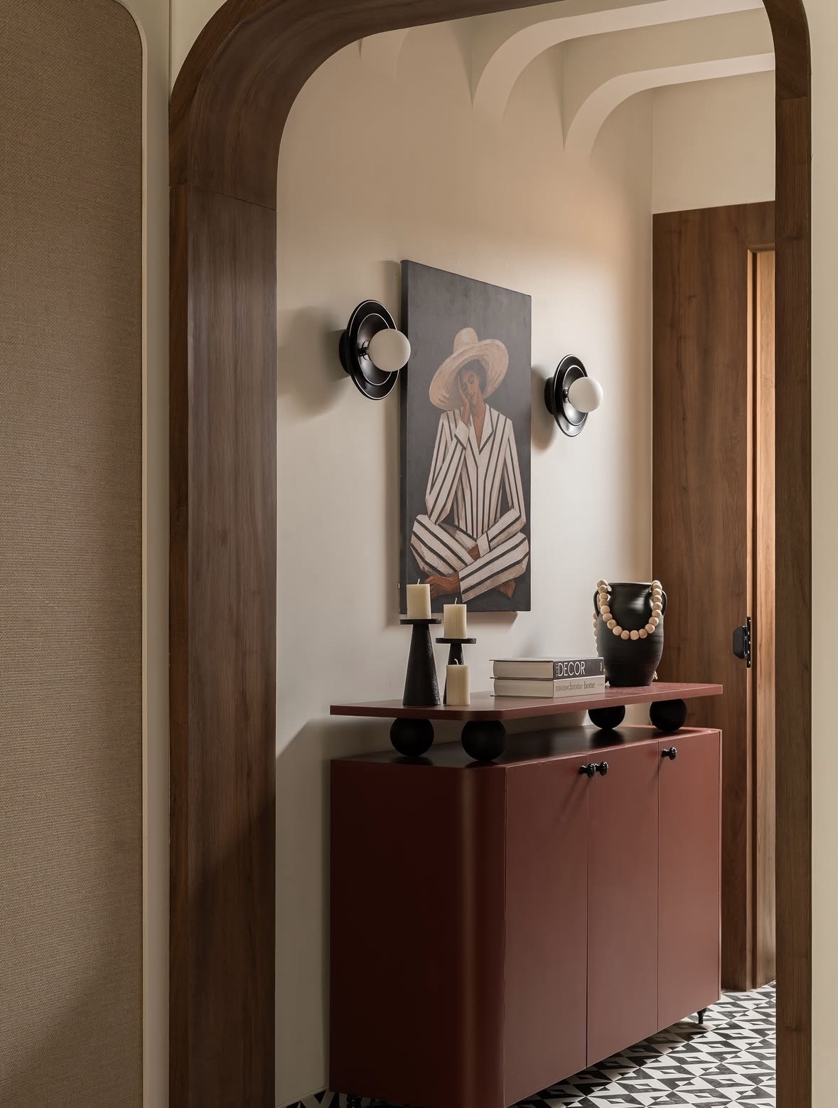 Timber arch opening onto a terracotta cabinet, a framed painting, two round wall lights and a checked black-and-white floor