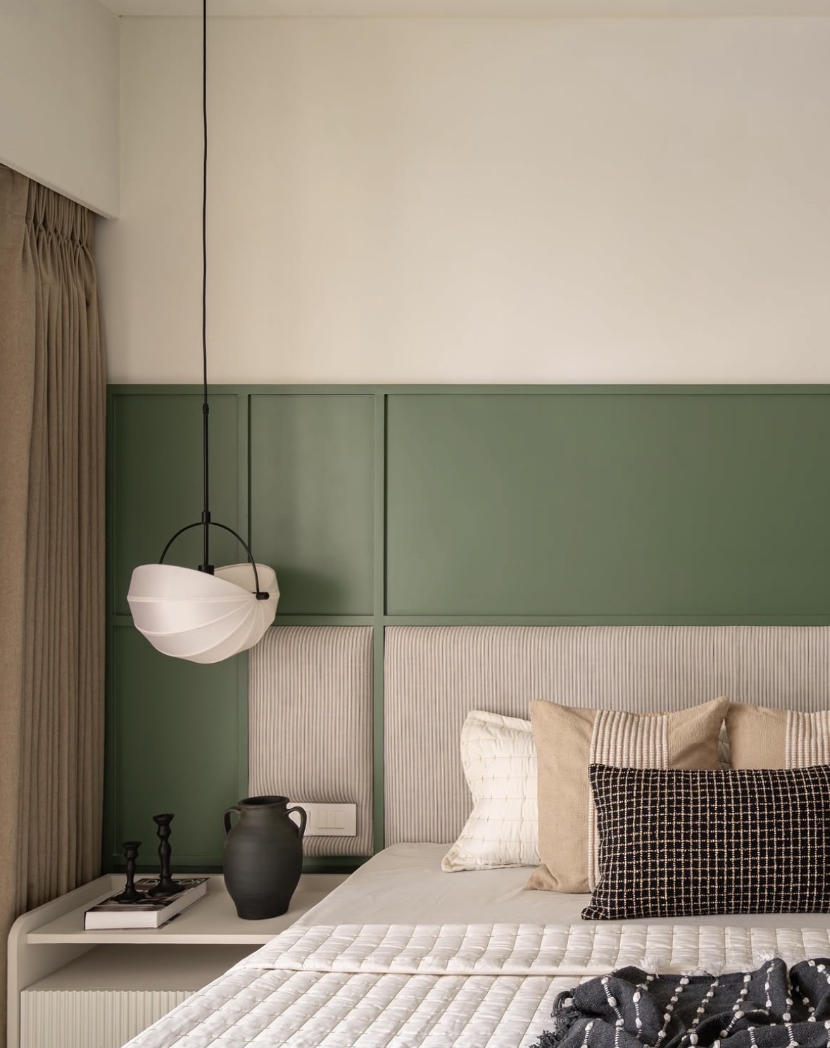 Bed with a striped upholstered headboard against green panelling, a paper pendant lamp and a black vase