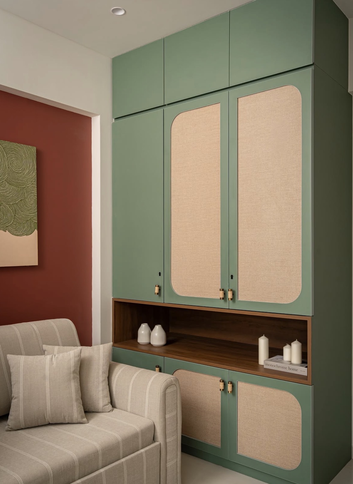 Sage-green wardrobe with rounded woven panels and an open timber shelf, beside a striped sofa and a terracotta wall