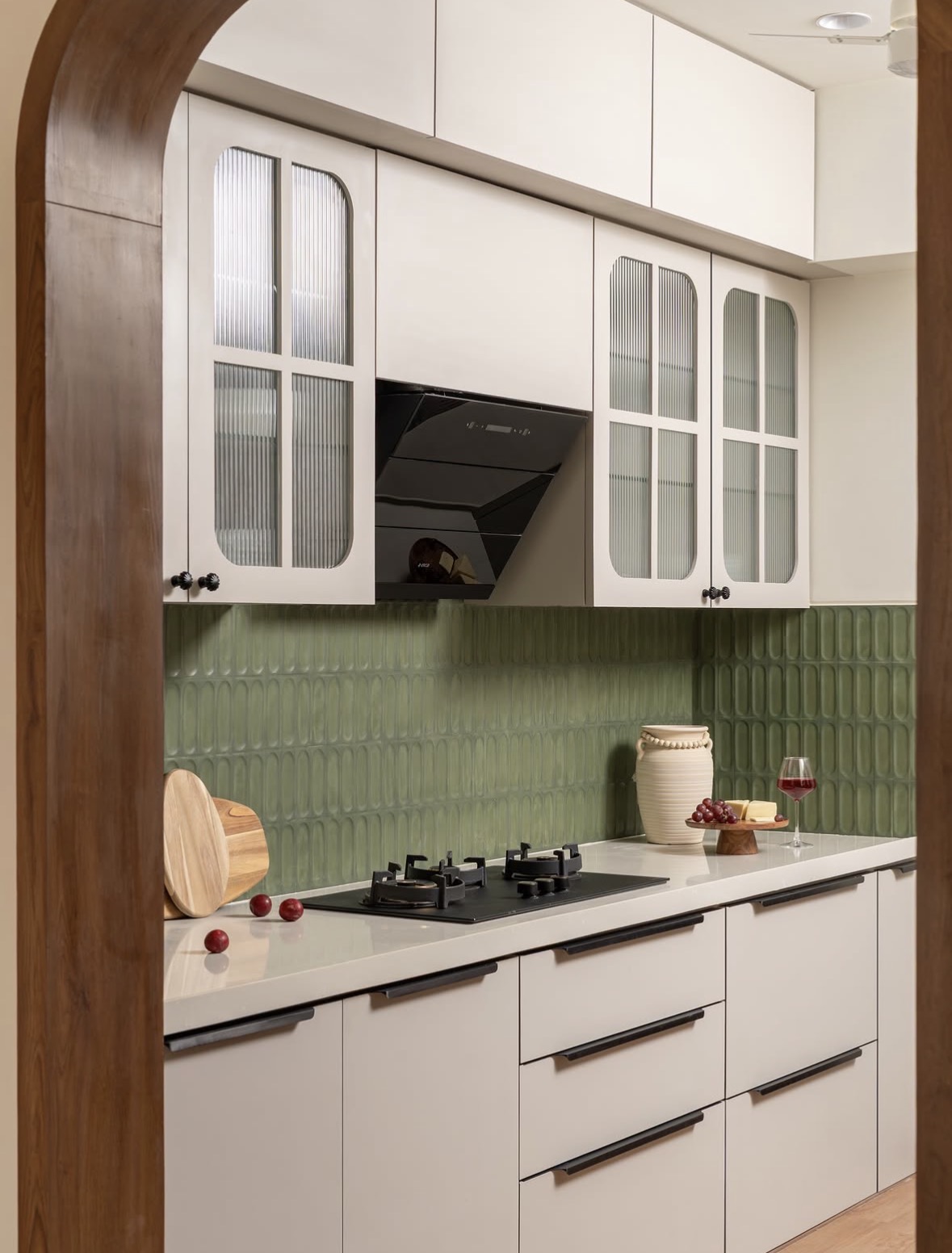 Kitchen with cream cabinets, reeded-glass doors and a sage-green tile backsplash beneath an arched timber doorway