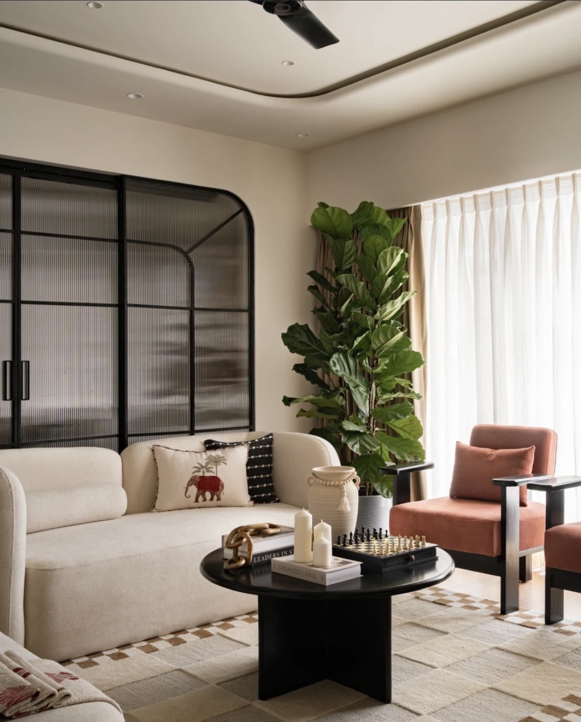 Rounded cream sofa with an embroidered elephant cushion, black-framed reeded-glass doors and a tall fiddle-leaf fig