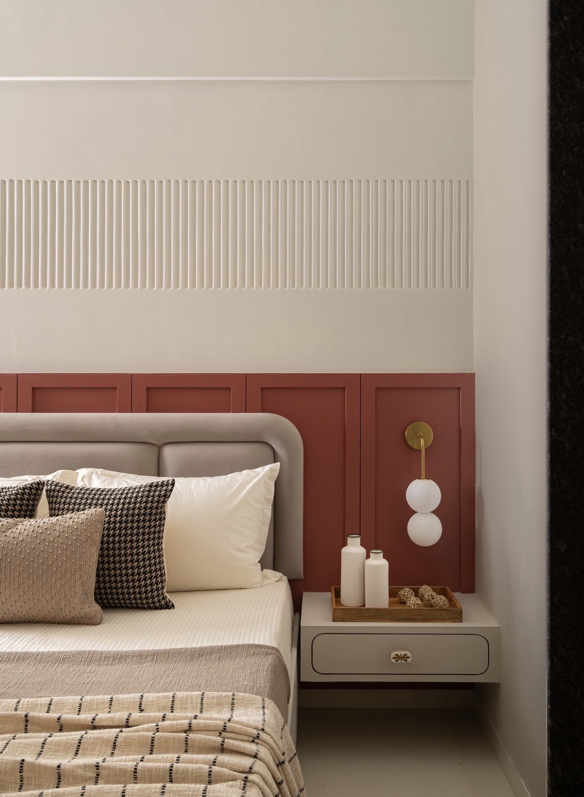 Bed with a rounded grey headboard against terracotta panelling, with a brass wall light and a ribbed plaster band above