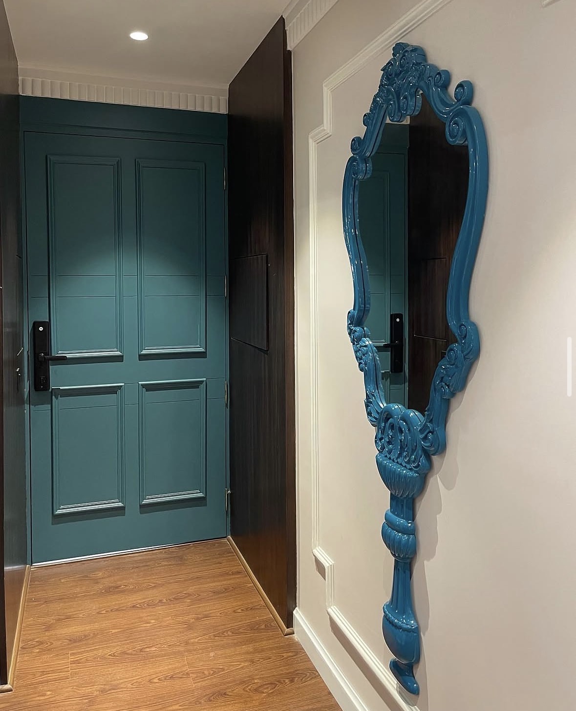 Teal panelled door beside a sculptural teal mirror in a hallway with dark wall panels and timber floor