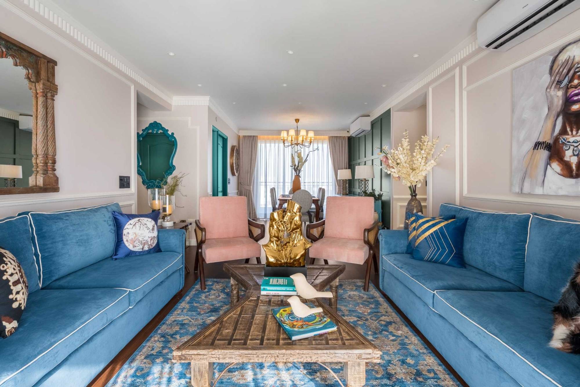 Living room with two cerulean velvet sofas, blush cane armchairs, a carved timber coffee table and a gold sculpture on a blue rug