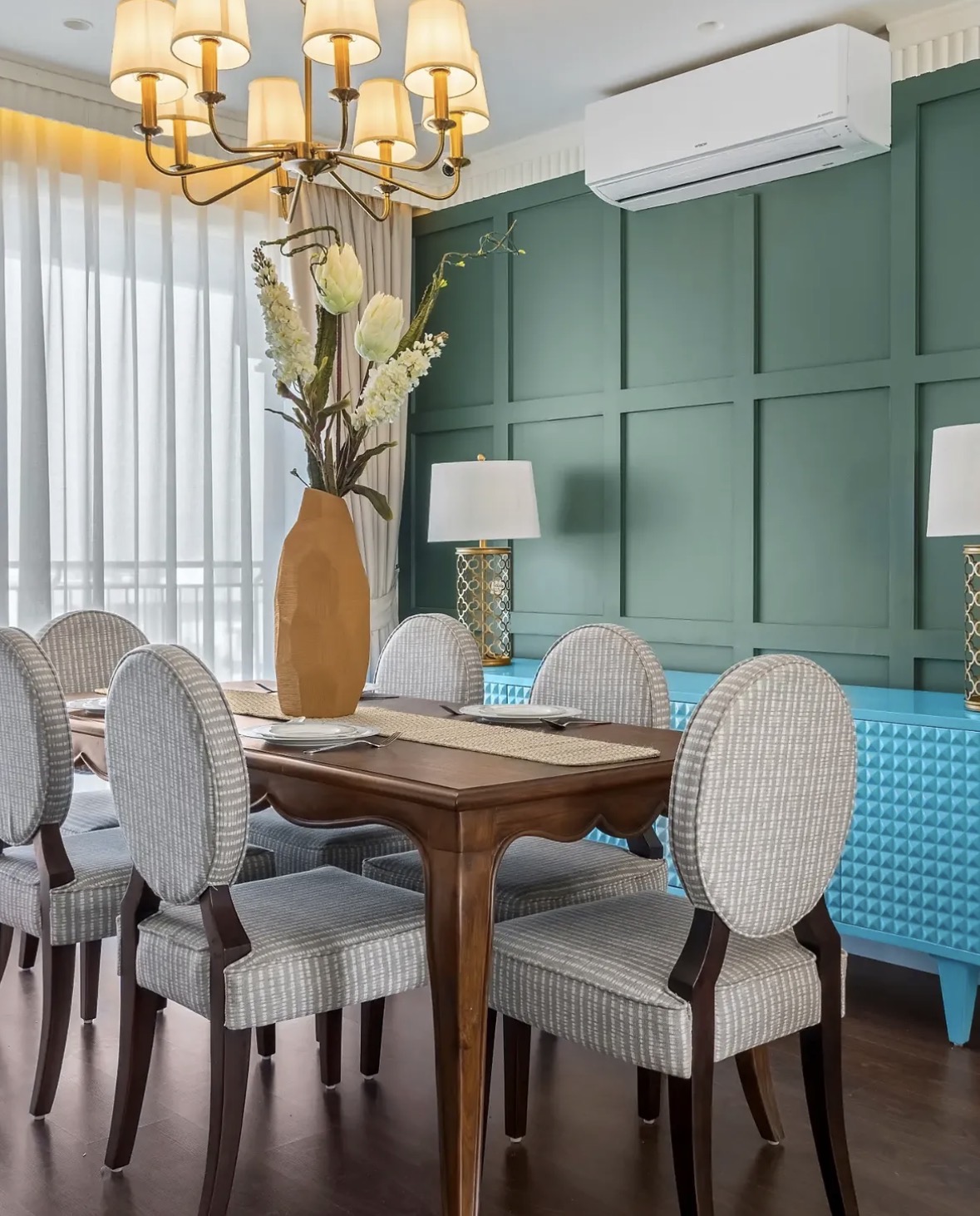 Dining table with oval-backed chairs in woven grey fabric, a brass chandelier and a green panelled wall