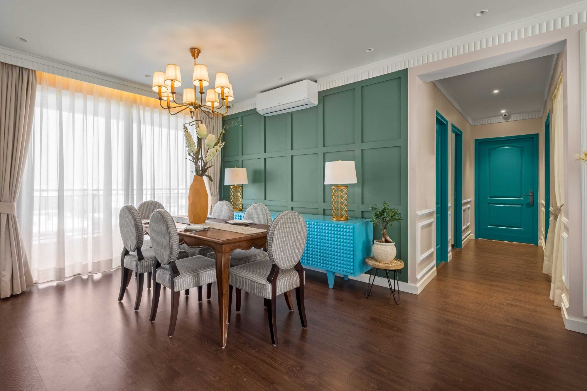 Dining table and chairs against a green panelled wall with a studded blue sideboard, leading to a hallway of teal doors