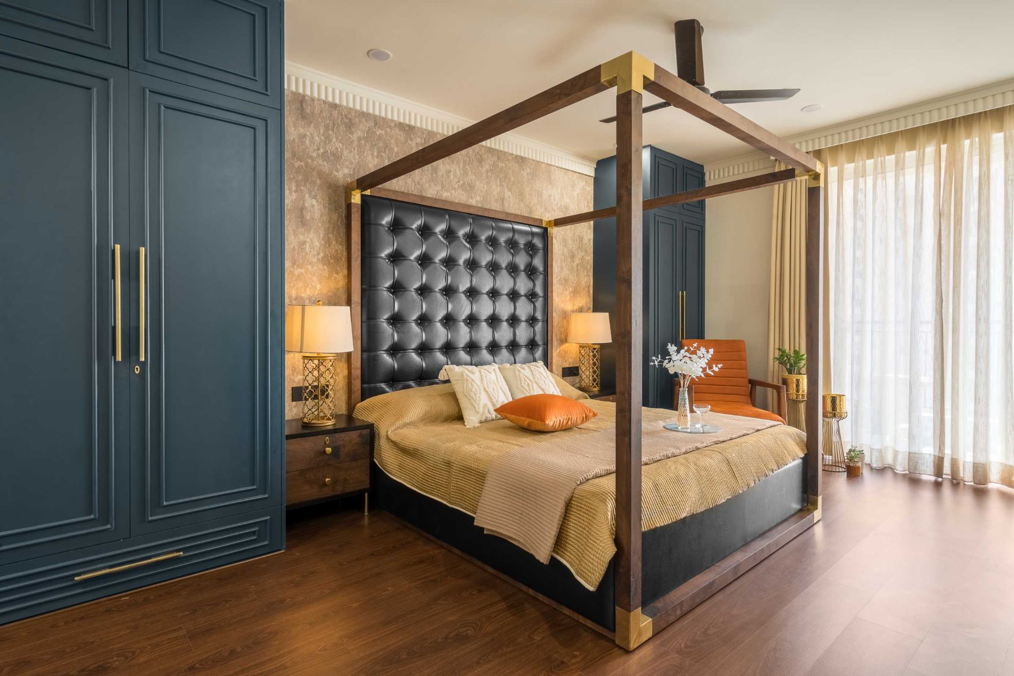Four-poster bed with a tall buttoned black headboard, brass corners, gold lamps and deep blue panelled wardrobes