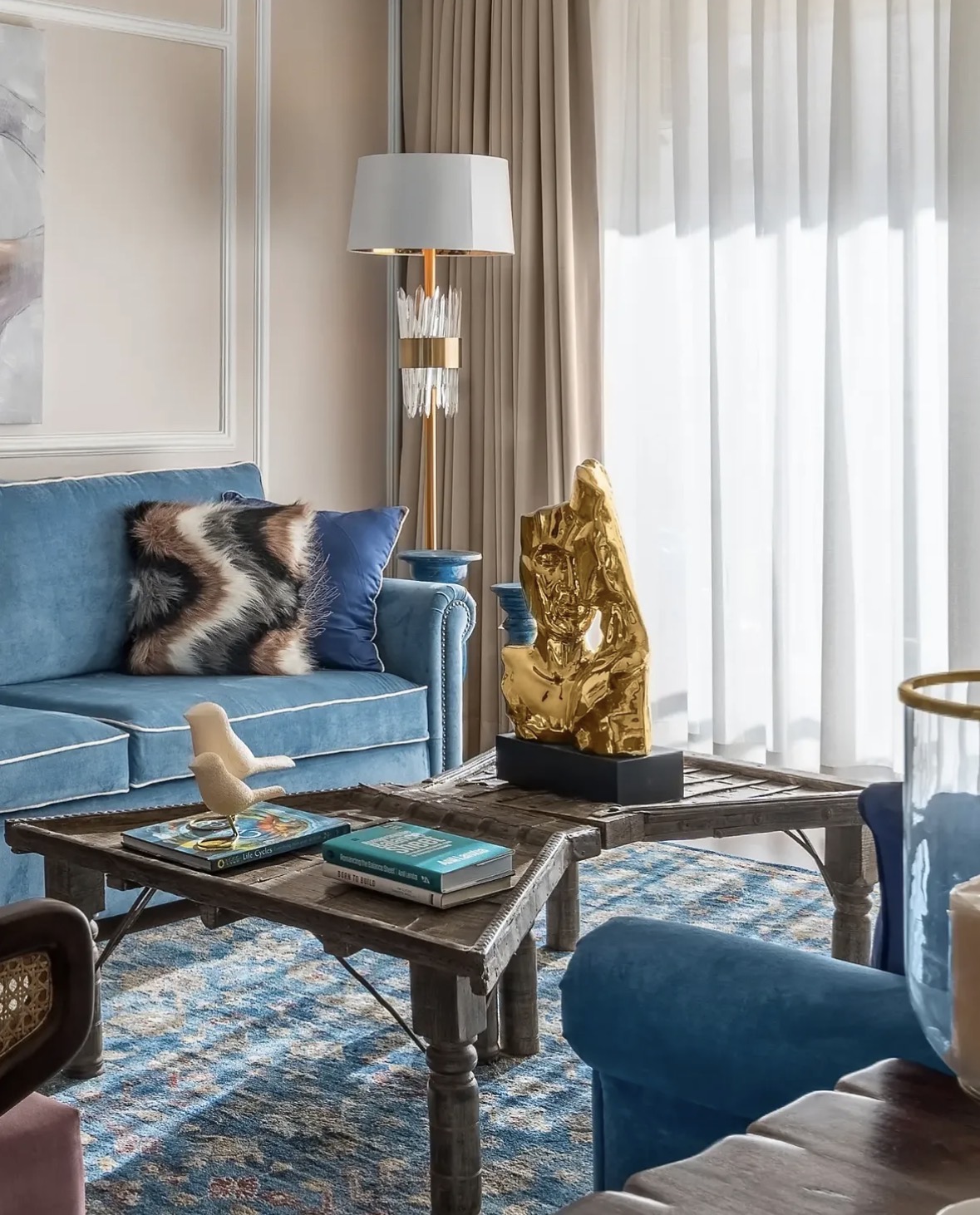 Gold sculpture and a pair of small books on a carved timber coffee table, with a blue sofa and a crystal floor lamp