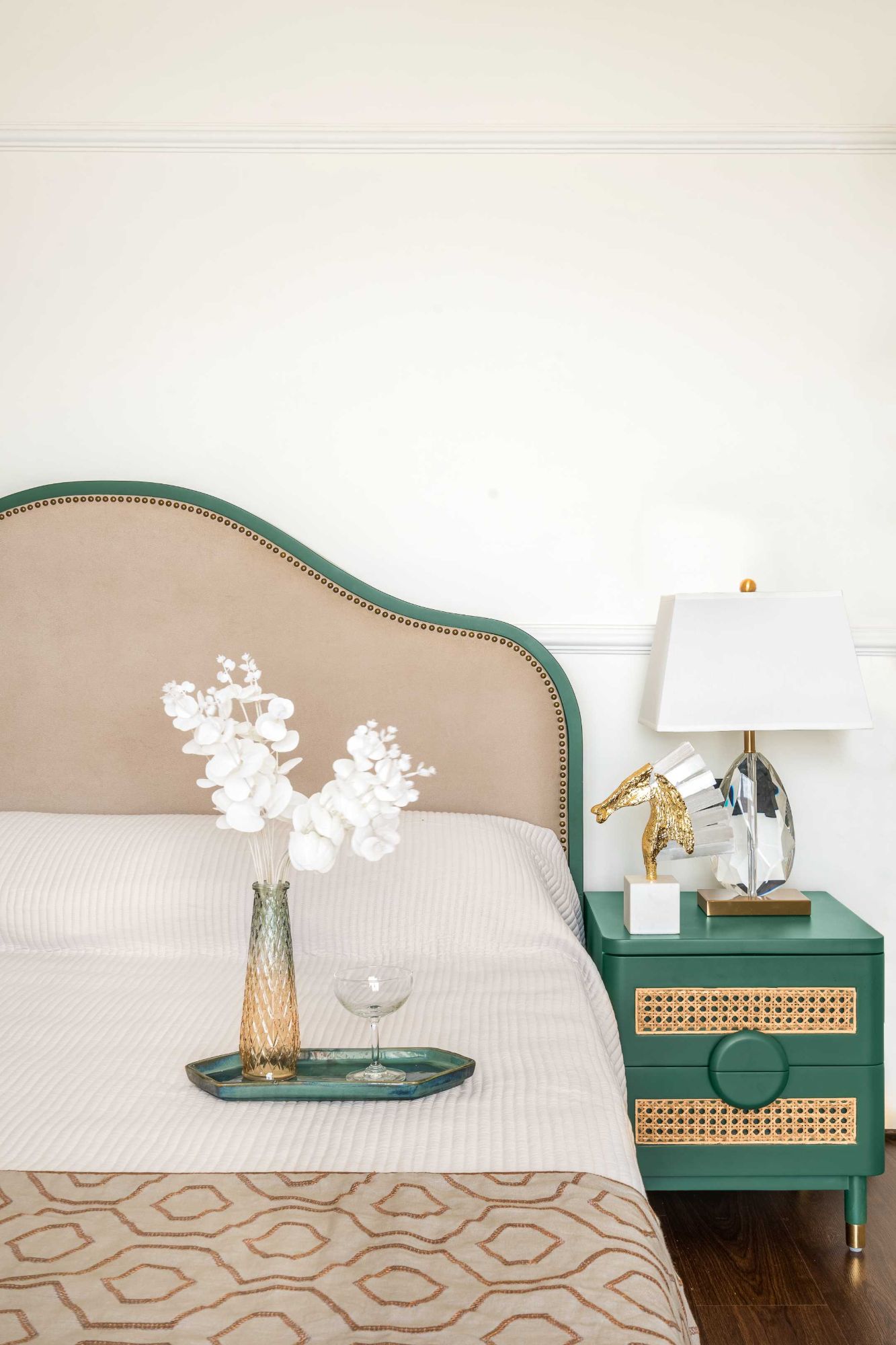 Bed with a curved green headboard studded in brass, and a green cane-fronted bedside with a crystal lamp