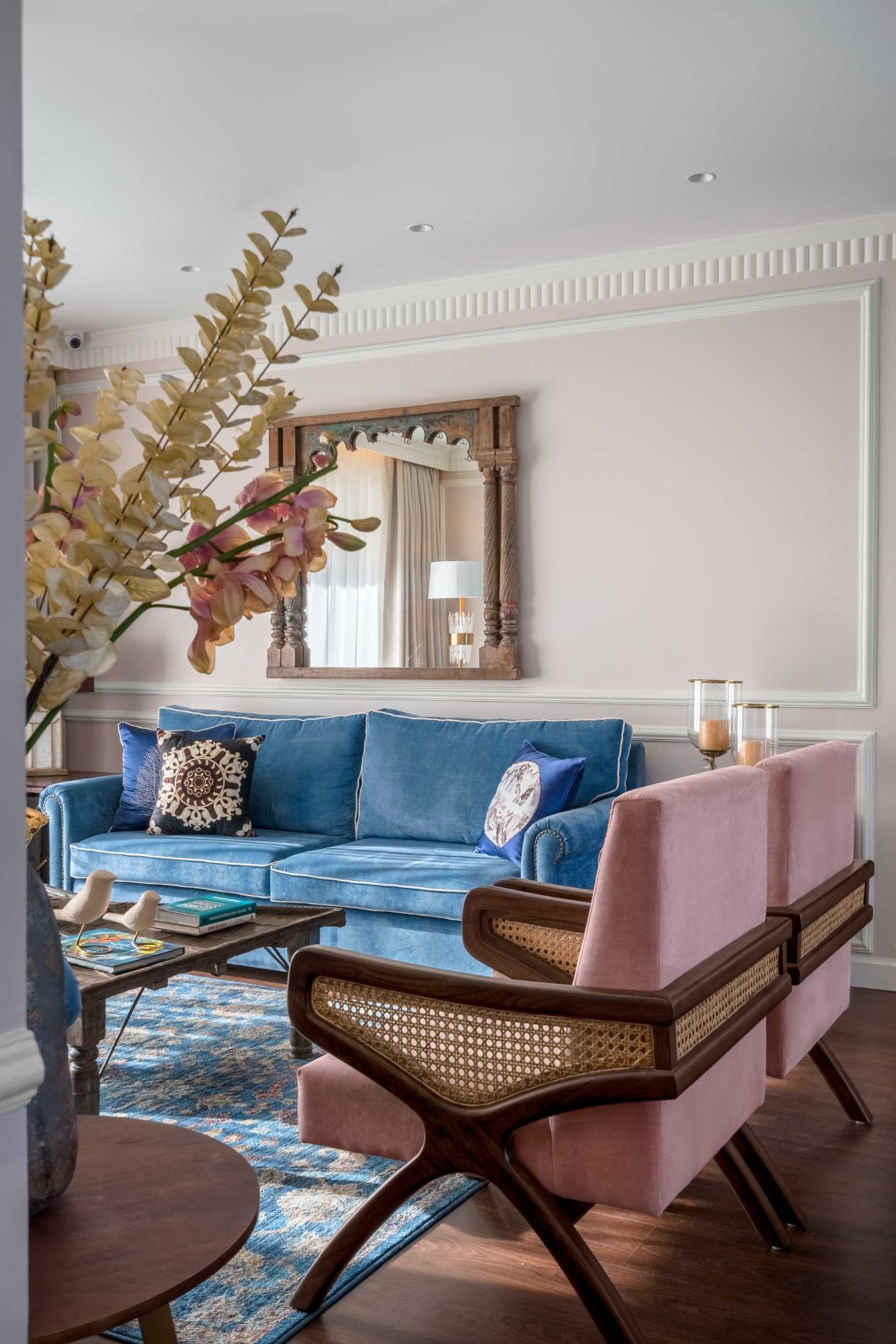 Blue sofa and a pair of blush cane armchairs beneath a carved timber mirror, with a large vase of blossom in the foreground