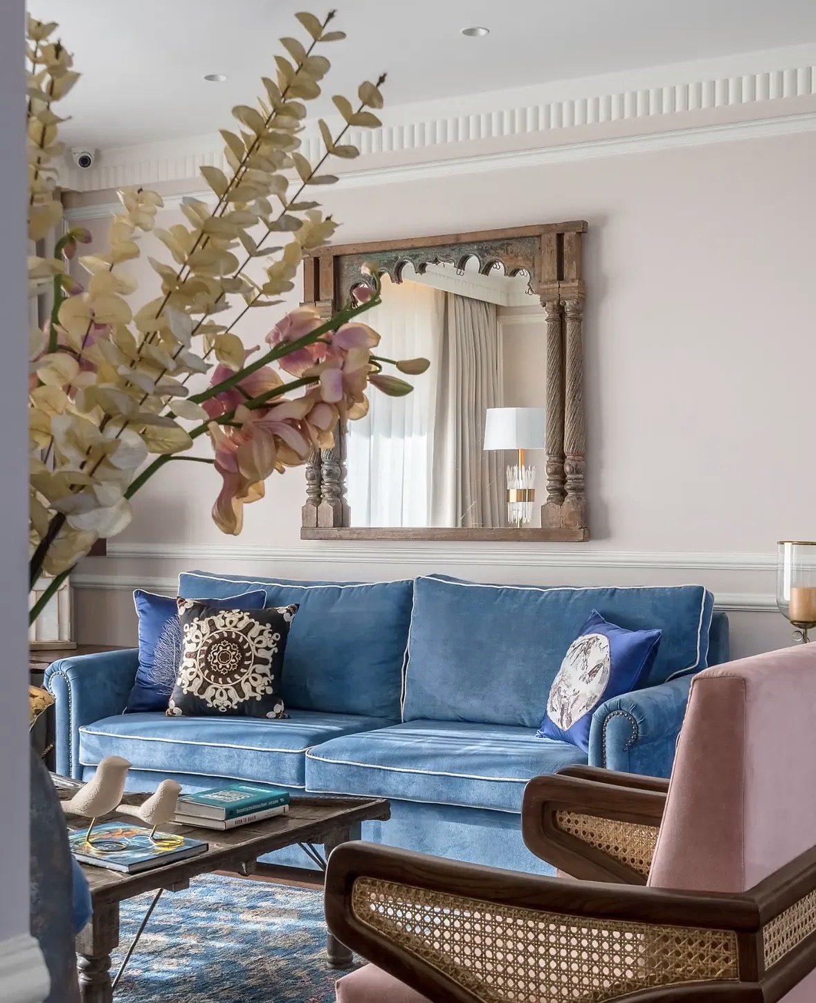 Cerulean velvet sofa with patterned cushions beneath a carved timber mirror, with a blush armchair in the foreground