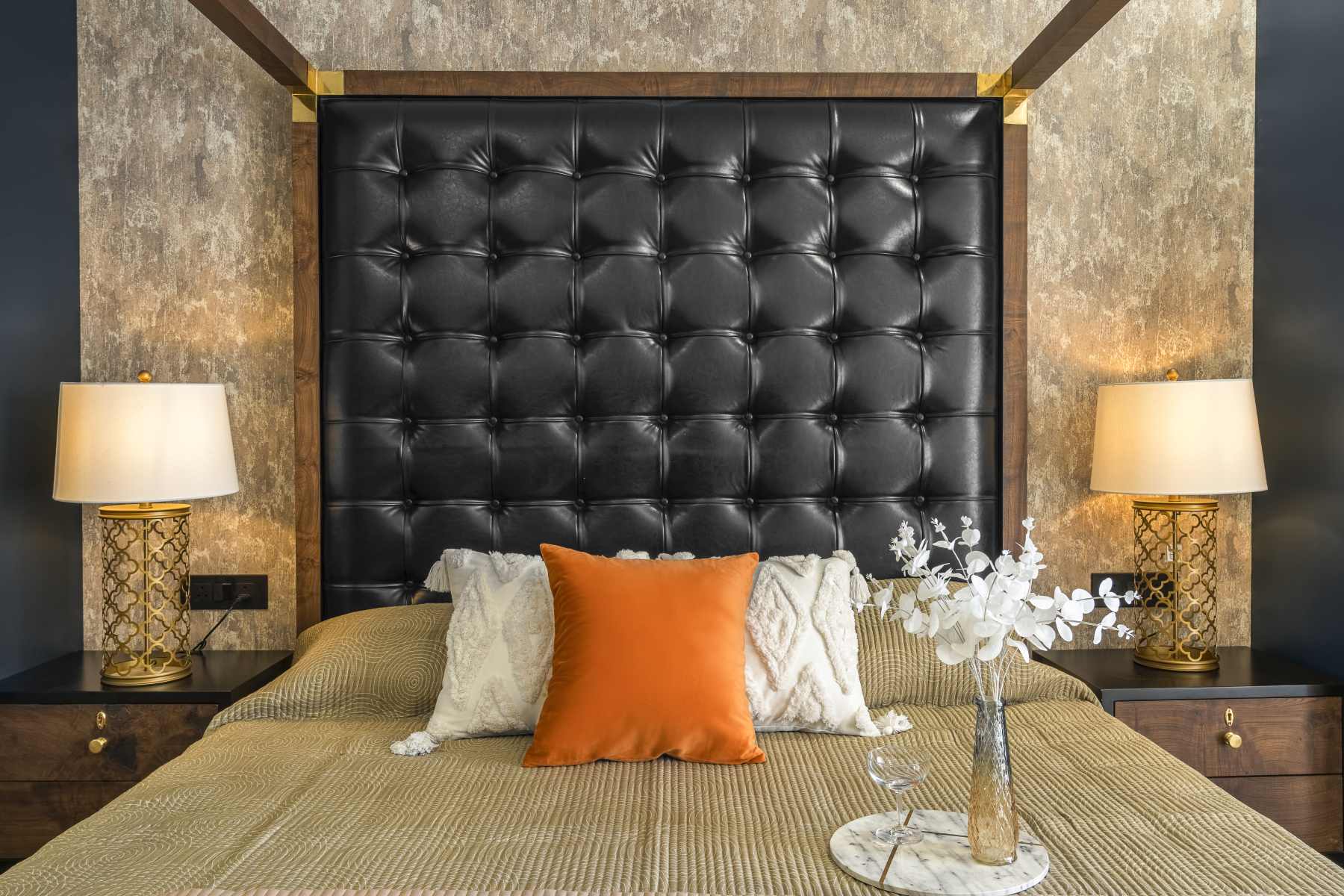 Buttoned black headboard with brass-capped frame, twin brass lamps, an orange cushion and a tray of flowers