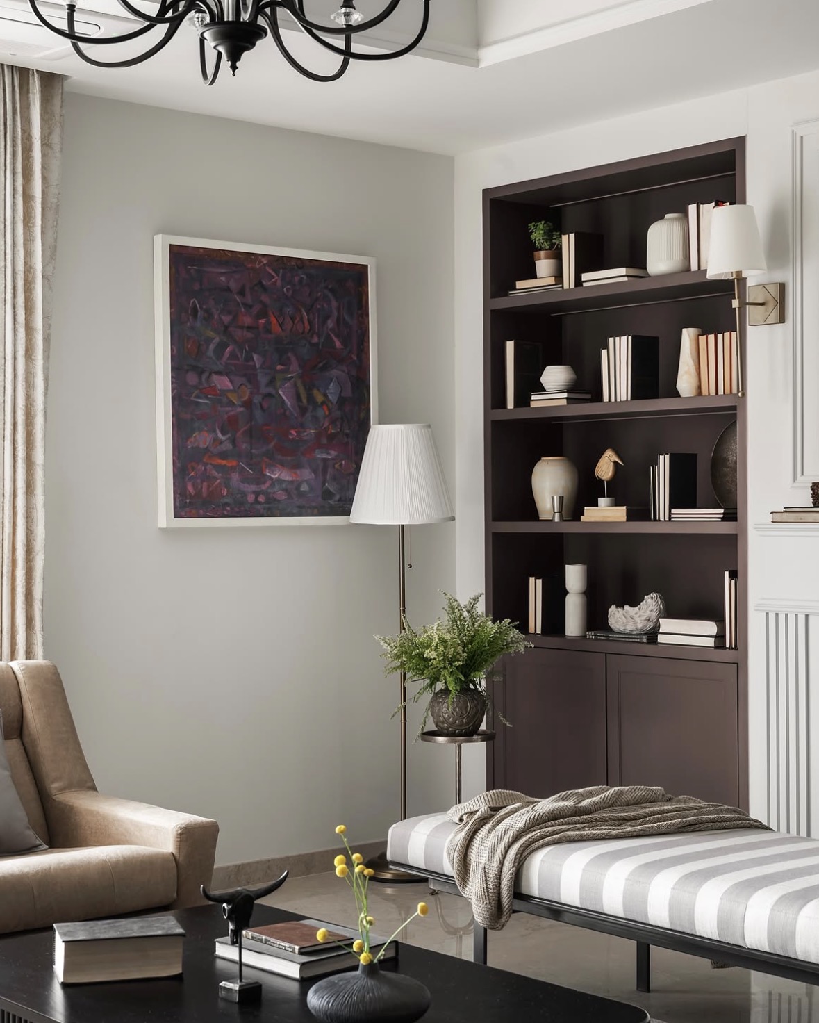Bronze-brown built-in shelves with books and ceramics, beside a framed painting and a sculptural console under a black chandelier