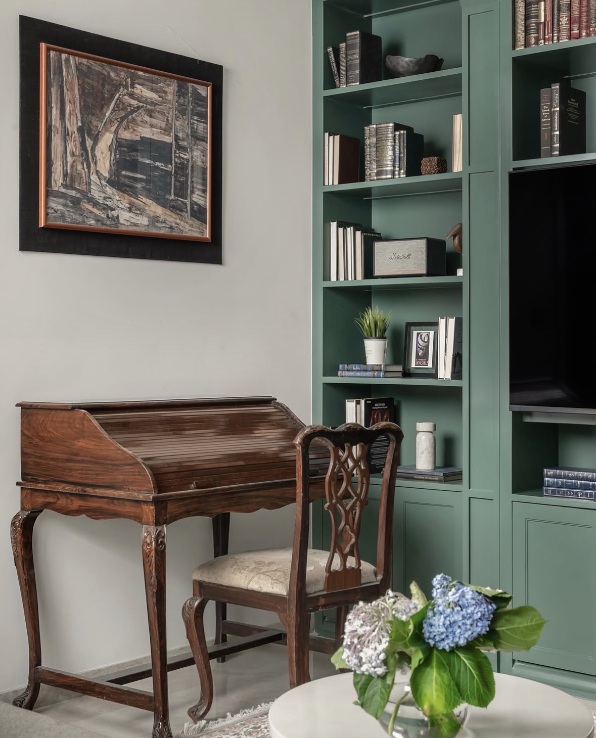 Antique rosewood writing desk and carved chair beside green shelves, under a framed painting