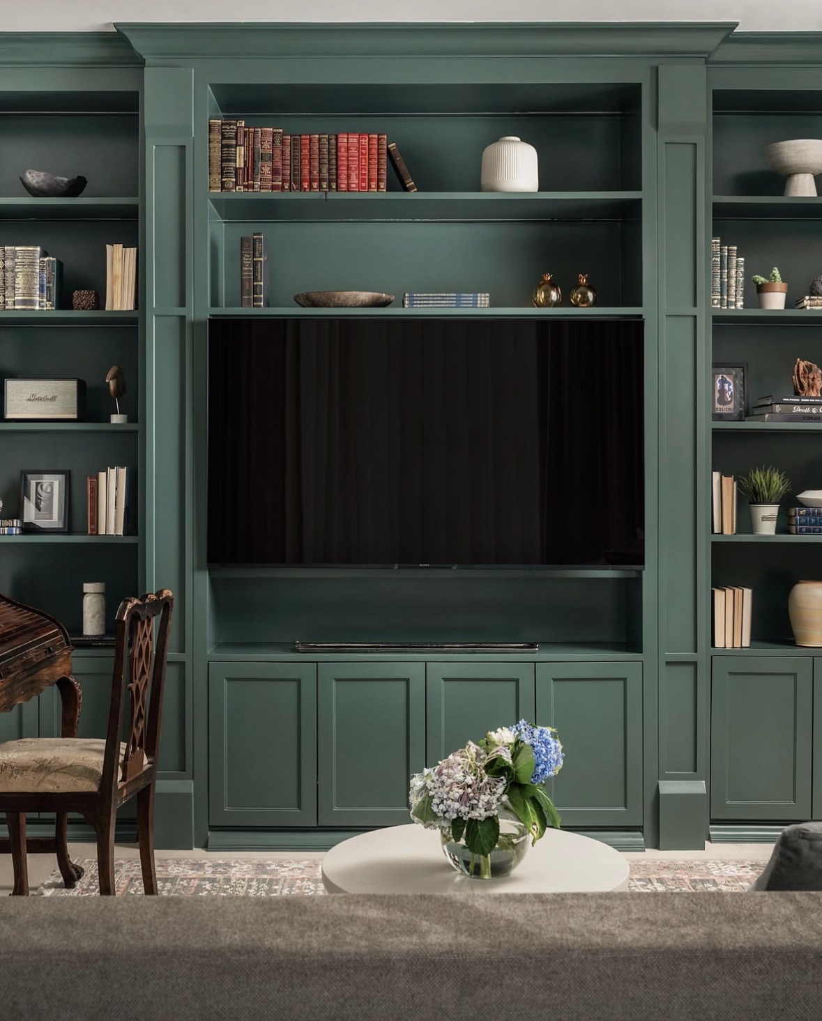 Forest-green built-in unit with a wall-mounted television, open shelves of books and objects, and a round table with hydrangeas