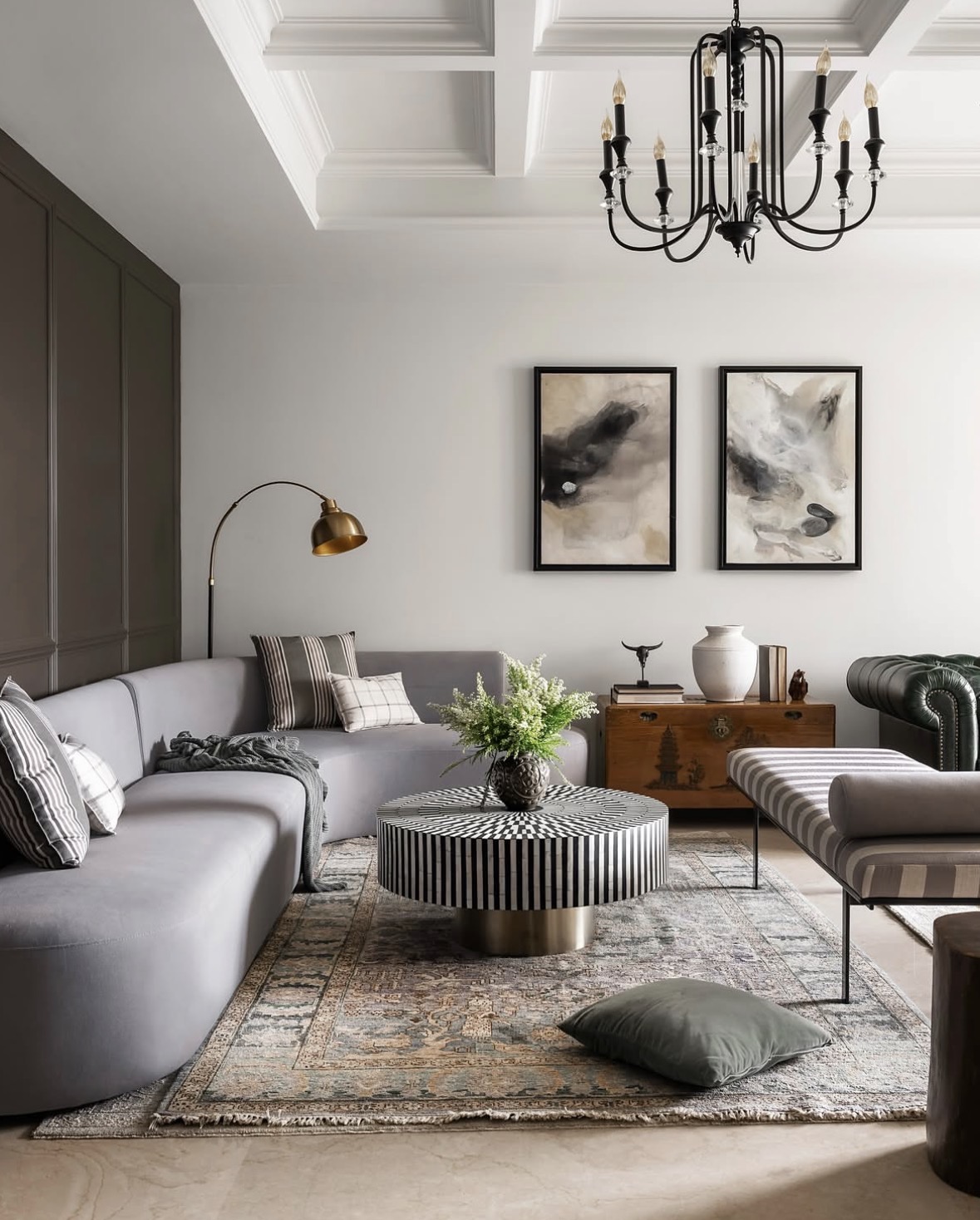 Curved grey sofa, a black-and-white striped round table, a brass arc lamp and two framed abstract paintings under a coffered ceiling