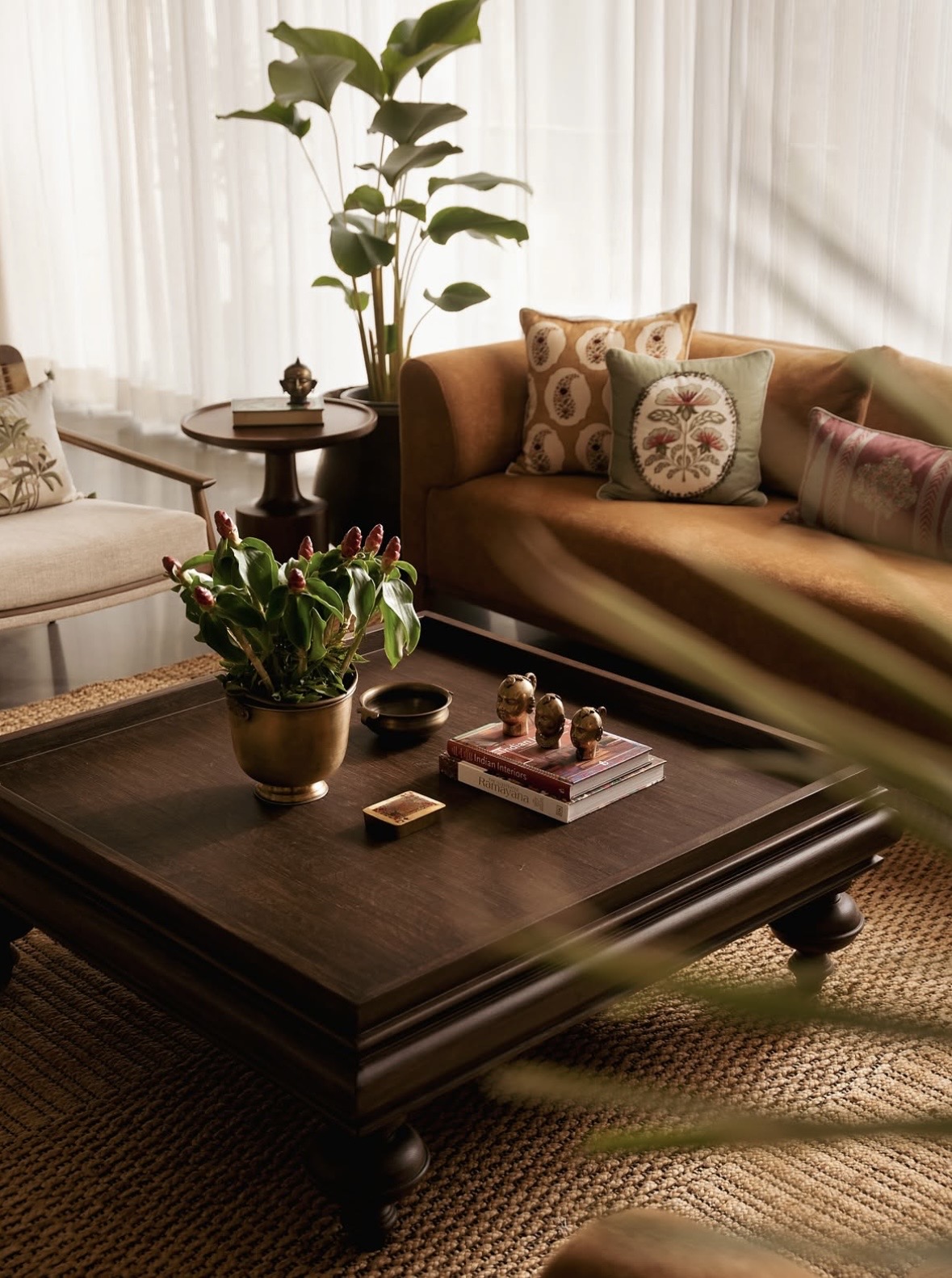 Brass pot of flowers and small brass heads on a dark carved coffee table, with an ochre sofa and a plant beyond