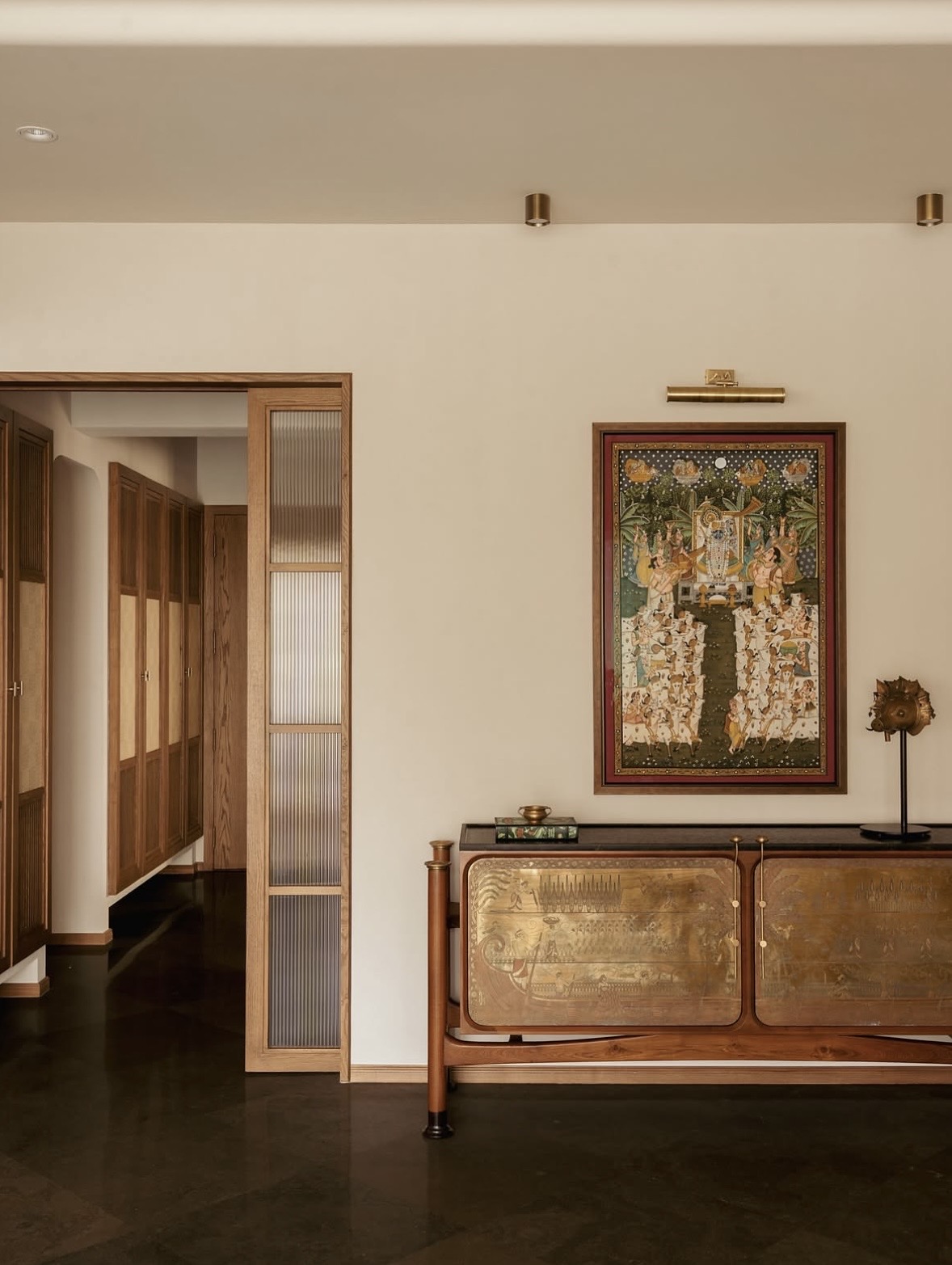 Brass-etched sideboard under a framed traditional painting and a brass picture light, beside a fluted-glass door