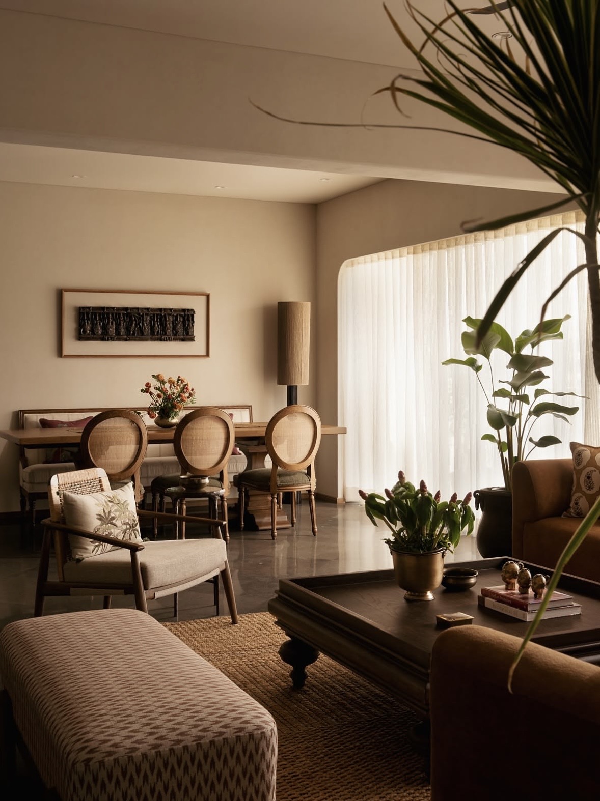 Dining table with cane-backed chairs and a long bench, opening onto a living area with a curved window of sheer curtains