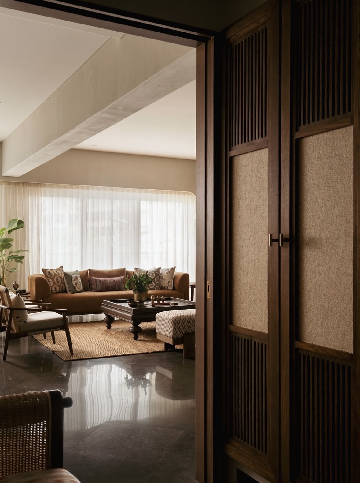 View through a timber doorway with slatted and woven panels into a living room with an ochre sofa and sheer curtains