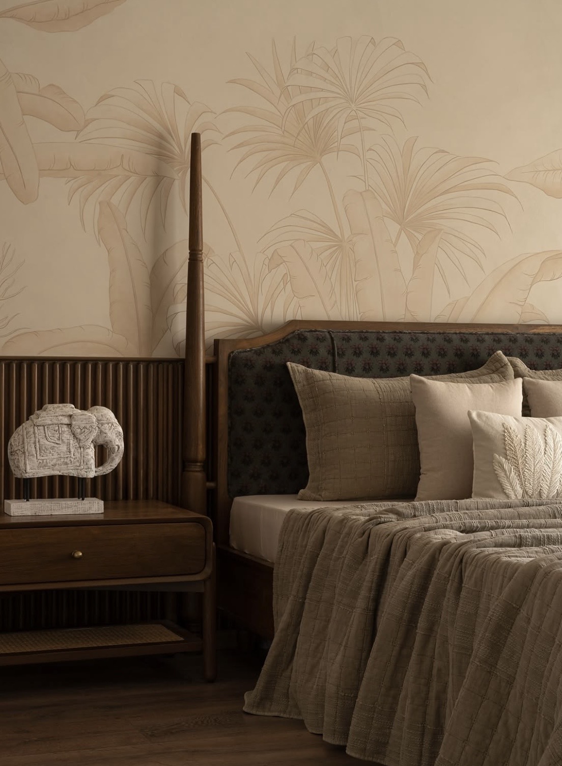 Bed against a palm-leaf mural with fluted timber panelling and a stone elephant on the bedside table