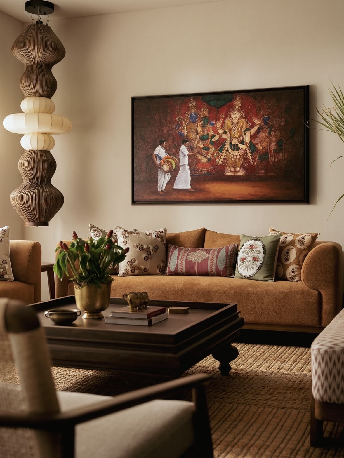 Ochre sofa with embroidered cushions beneath a large framed painting, a ribbed pendant lamp and a low carved timber table