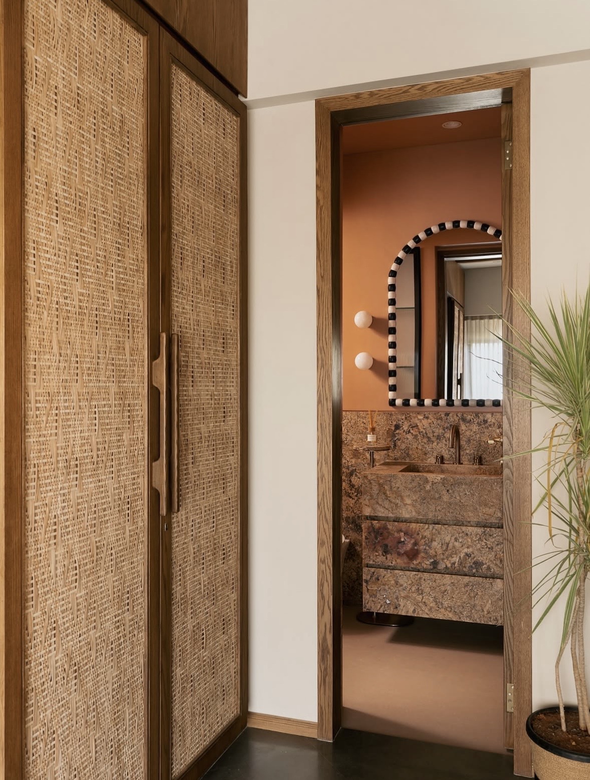 Woven wardrobe doors beside a timber doorway leading to a powder room with terracotta walls and an arched beaded mirror