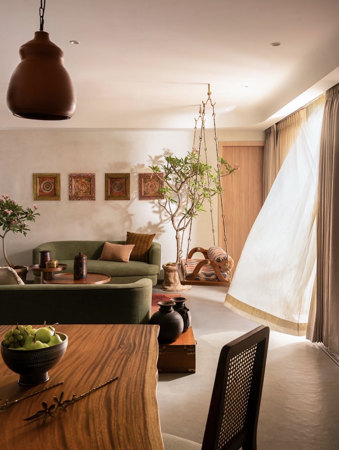 Green sofa, a carved swing by a billowing sheer curtain and a flowering tree, seen past a timber dining table