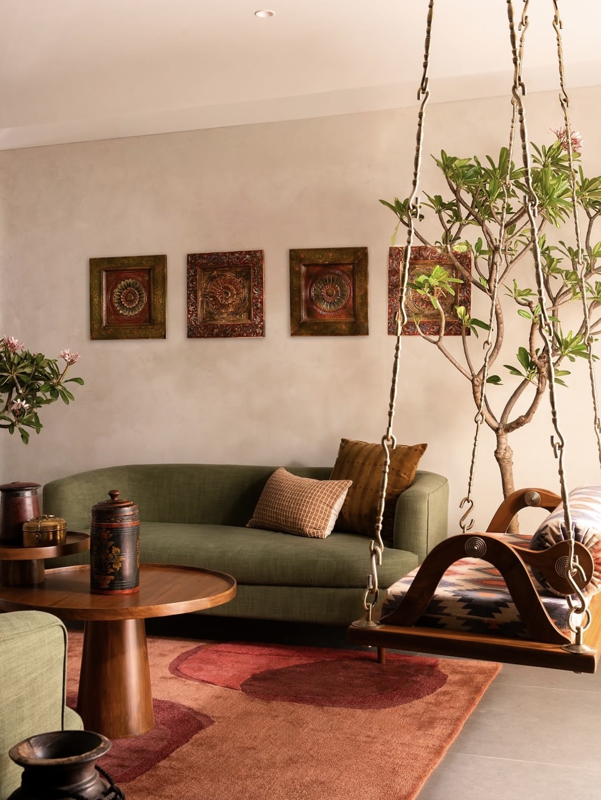 Olive sofa below four carved wall plaques, with a swing and a flowering tree to the right