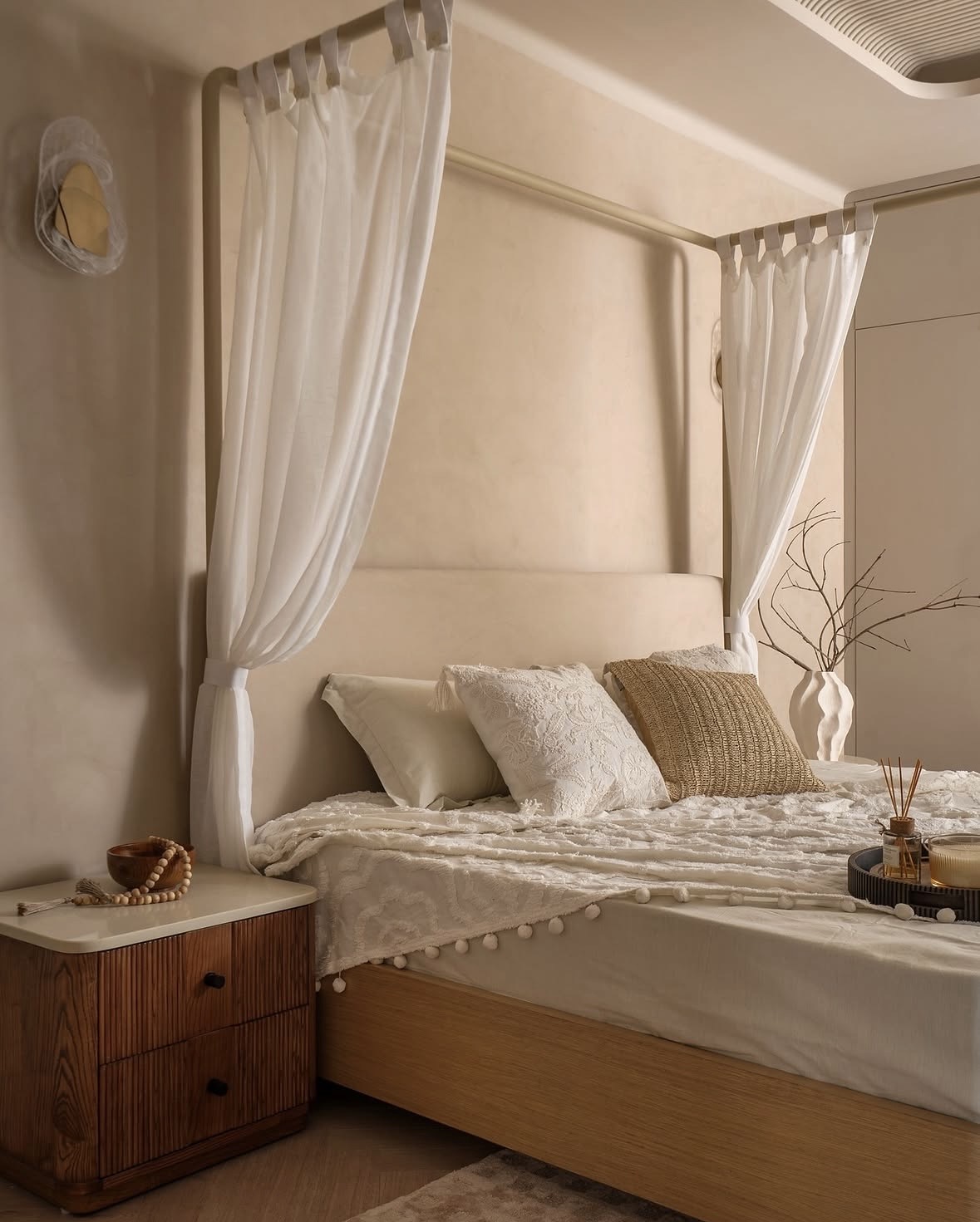Four-poster bed with white drapes, a cream headboard, textured bedding and a fluted timber bedside table