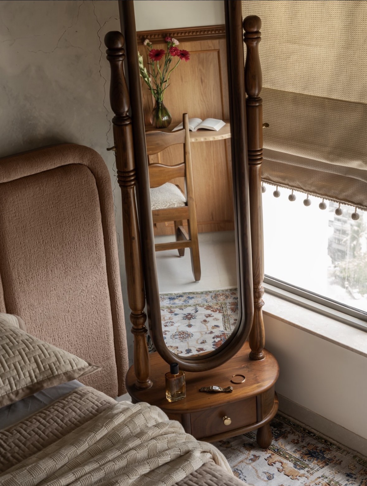 Wooden cheval mirror on a turned stand beside a brown textured headboard, with a window, a roman blind and a patterned rug