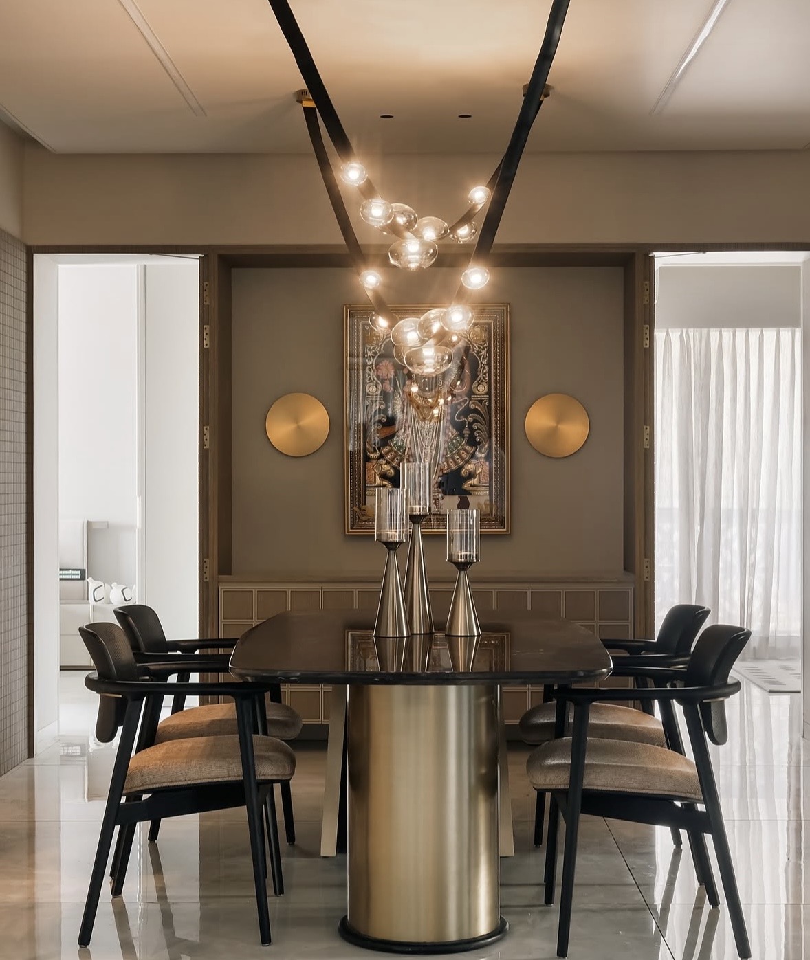 Dining table with a brass base, black chairs, brass wall discs and a framed painting under a ribbon of glass globes