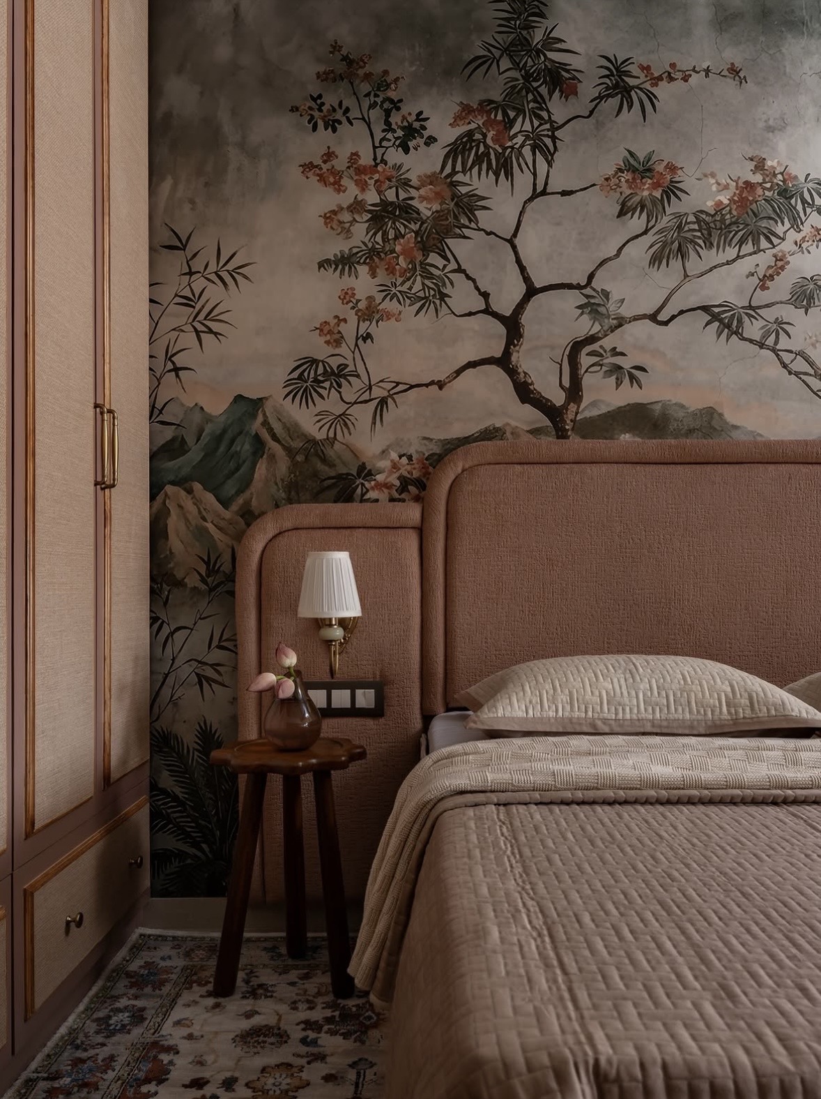 Bed with a rose upholstered headboard against a hand-painted landscape mural, beside a fabric-panelled wardrobe