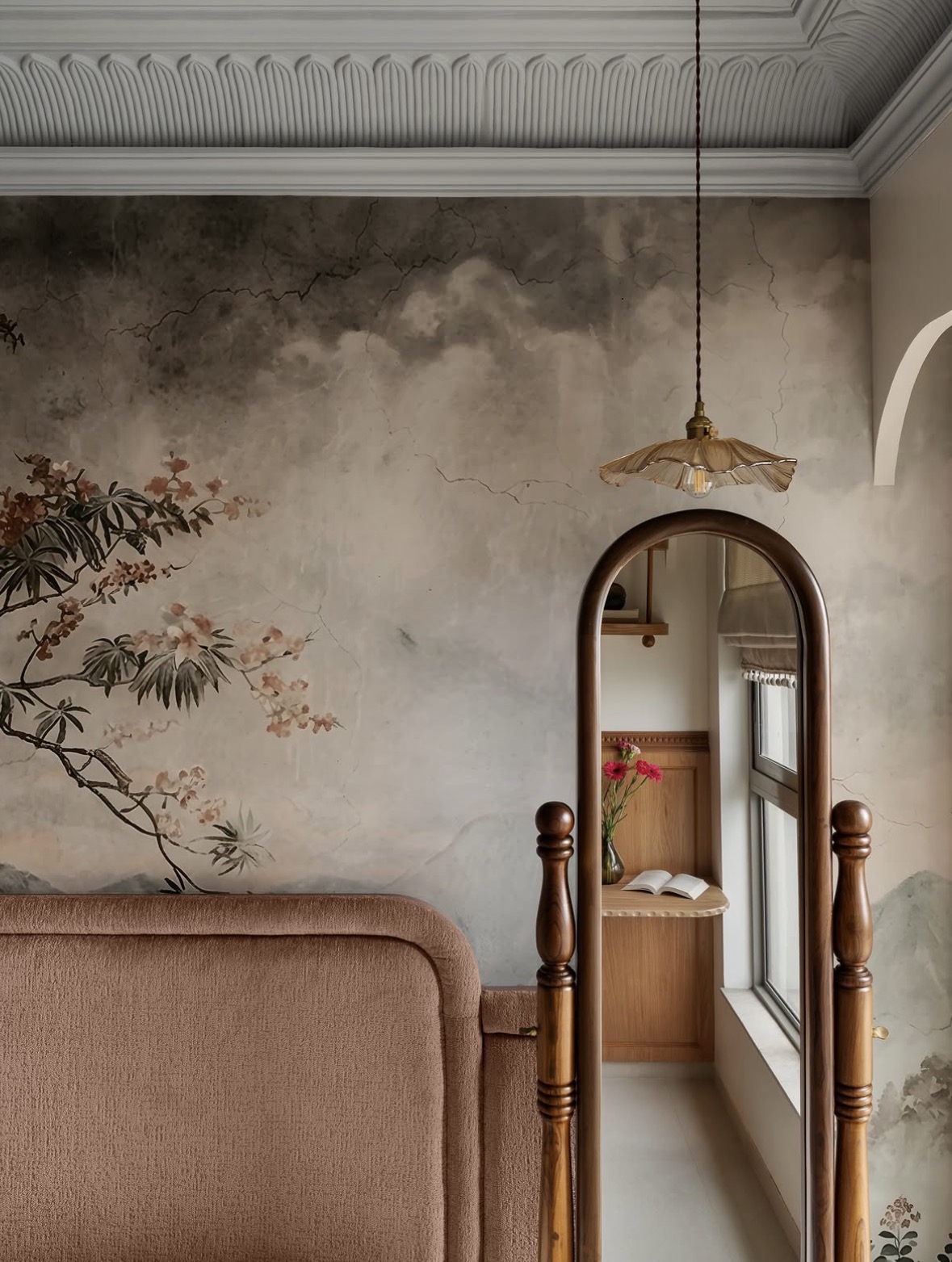 Arched wooden mirror in front of a painted landscape mural, with a scalloped pendant lamp, a plaster cornice and an arched niche