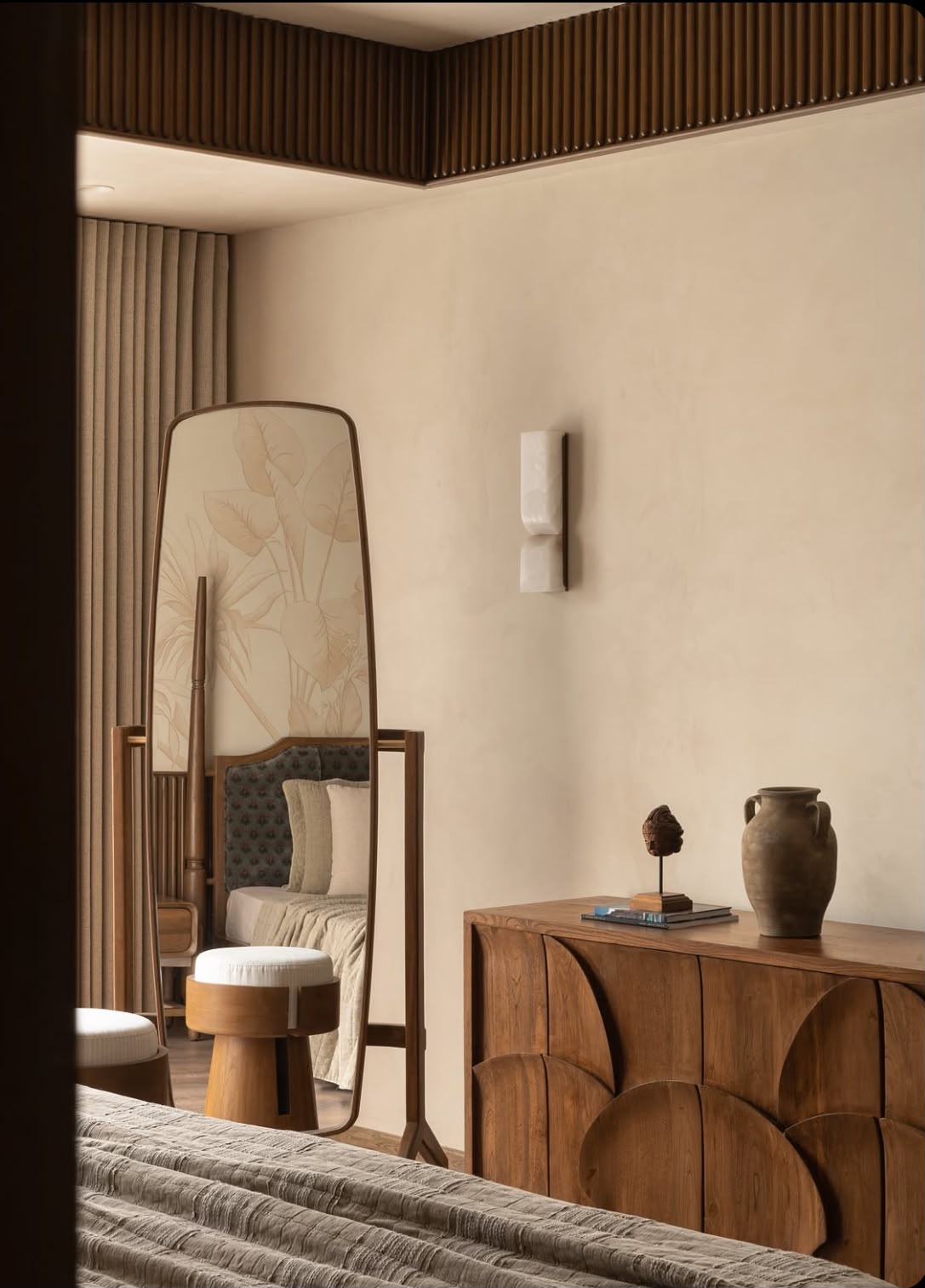 Bedroom corner with a slatted timber ceiling edge, a wall light, a tall mirror and a carved wooden sideboard